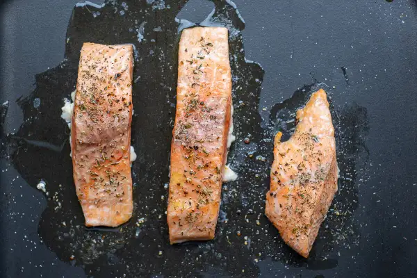 Drei saftige Lachsfilets mit Kräutern werden im Ofen auf einer schwarzen Pfanne zubereitet.