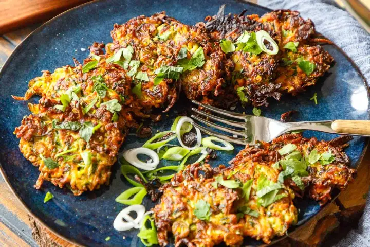Knusprige Zucchini-Puffer, garniert mit frischen Kräutern und Frühlingszwiebeln, liegen auf einem blauen Teller. Eine Gabel liegt daneben.